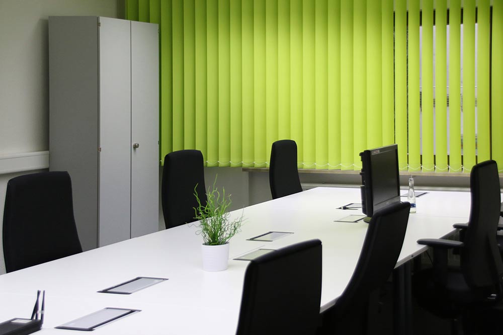 Konferenzraum mit langem weißen Tisch, davor Schreibtischstühle. Im Hintergrund ein grüner Vorhang